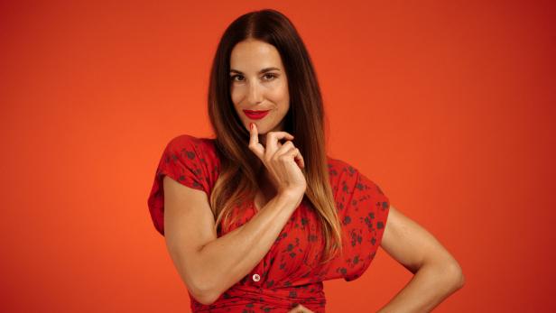 Eine Frau in einem roten Kleid mit Blumenmuster posiert vor einem orangefarbenen Hintergrund.