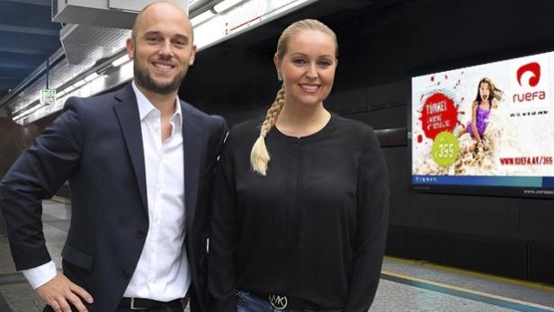 Ein Mann im Anzug und eine Frau stehen vor einer Werbung für „ruefa Reisen“ in einer U-Bahn-Station.