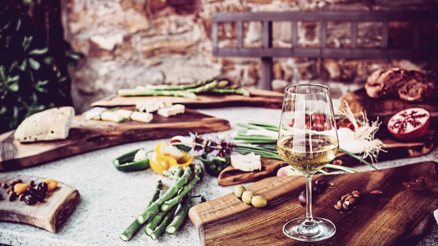 Ein Tisch mit einem Glas Weißwein, Käse, Brot, Spargel und anderen Speisen angerichtet.
