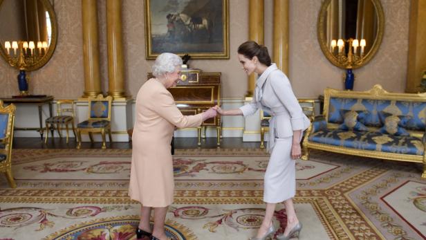 Königin Elisabeth II. und Angelina Jolie geben sich die Hand in einem opulenten Raum.