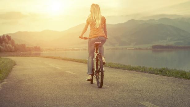 Eine Frau fährt mit dem Fahrrad auf einer Straße in Richtung eines Sees und der Berge.