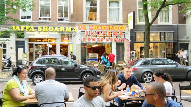 Menschen sitzen vor Restaurants in einer belebten Straße und essen.
