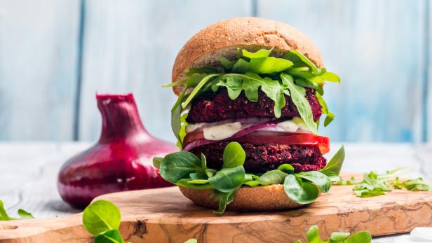 Ein doppelter Rote-Bete-Burger mit Salat, Tomaten und Zwiebeln auf einem Holzbrett.