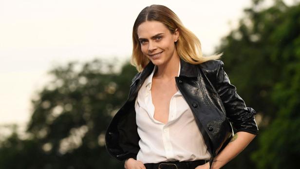 Cara Delevingne posiert in einer schwarzen Lederjacke und einer weißen Bluse.