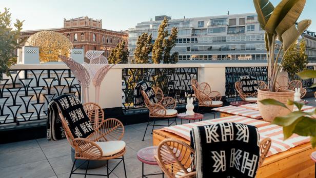 Eine Dachterrasse mit Korbstühlen, Tischen und Blick auf Gebäude in der Stadt.