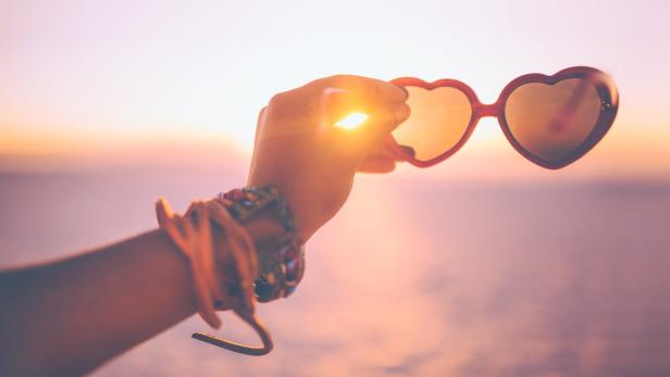 Eine Hand hält eine herzförmige Sonnenbrille vor einem Sonnenuntergang.