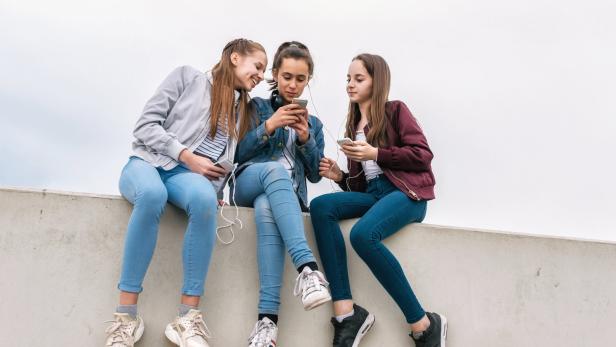 Drei Teenager sitzen auf einer Mauer und schauen auf ihre Handys.