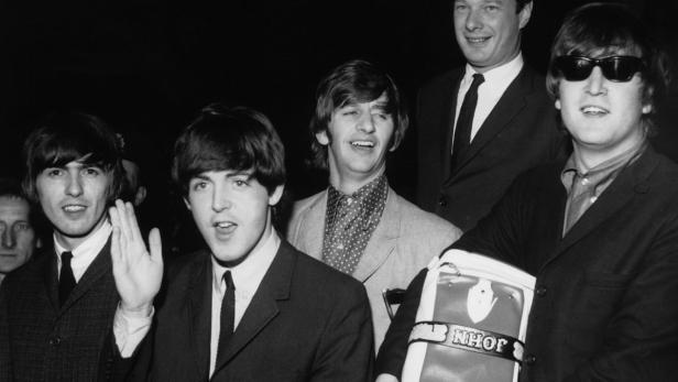 Die Beatles posieren mit einer Tasche mit der Aufschrift „John“ für ein Schwarzweißfoto.