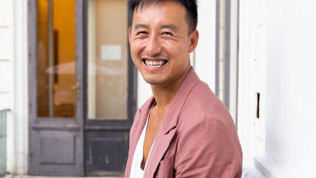 Ein lächelnder Mann mit Blazer lehnt an einer weißen Wand.