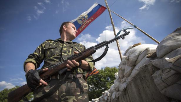 Ein Soldat in Tarnkleidung steht mit einem Gewehr vor Sandsäcken und einer russischen Flagge.