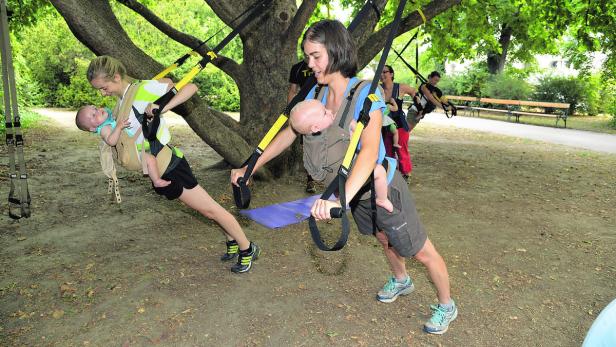 Zwei Mütter trainieren mit ihren Babys in Tragetüchern an einem Baum mit TRX-Bändern.