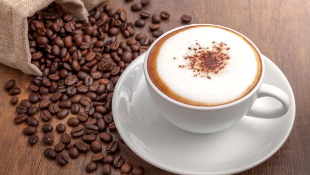 Eine Tasse Cappuccino steht auf einer Untertasse neben einem Sack Kaffeebohnen.