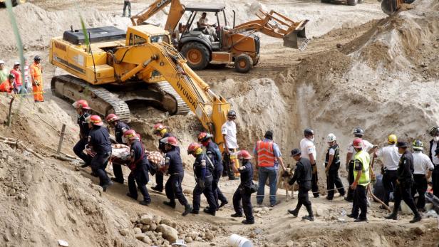 Rettungskräfte tragen eine Person auf einer Trage an einer Baustelle vorbei, im Hintergrund Bagger.
