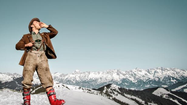 Ein Mann in Tracht steht auf einem schneebedeckten Berg und ruft.