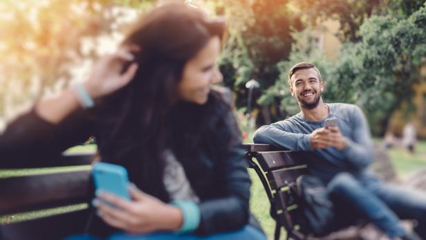 Ein lächelnder Mann sitzt mit seinem Handy auf einer Parkbank, während eine Frau neben ihm sitzt.