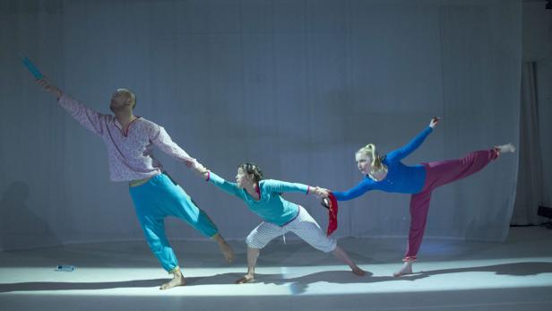 Drei Tänzer in farbenfrohen Kostümen führen eine synchrone Bewegung auf einer Bühne auf.