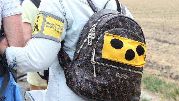 Person mit Rucksack und gelbem Armband mit Symbolen, im Freien auf einem Feldweg unterwegs.