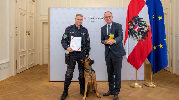 Ein Polizist mit Urkunde und sein Diensthund posieren mit einem Mann im Anzug vor der österreichischen und EU-Flagge.