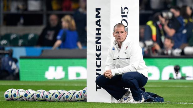 Ein Mann kauert neben einer Reihe von Rugbybällen vor einer Werbetafel.