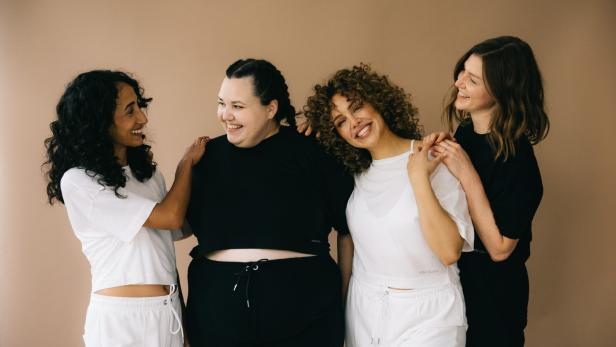 Vier Frauen unterschiedlicher Größe und ethnischer Zugehörigkeit stehen zusammen und lächeln.