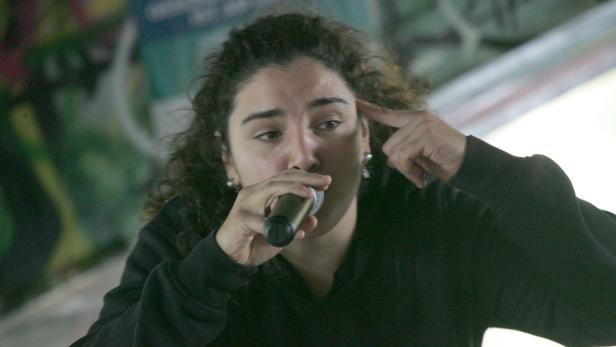 Eine Frau mit lockigem Haar rappt in ein Mikrofon und tippt sich an die Stirn.