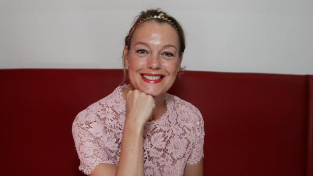 Eine lächelnde Frau mit rotem Lippenstift und einem Spitzenoberteil posiert vor einer roten Wand.