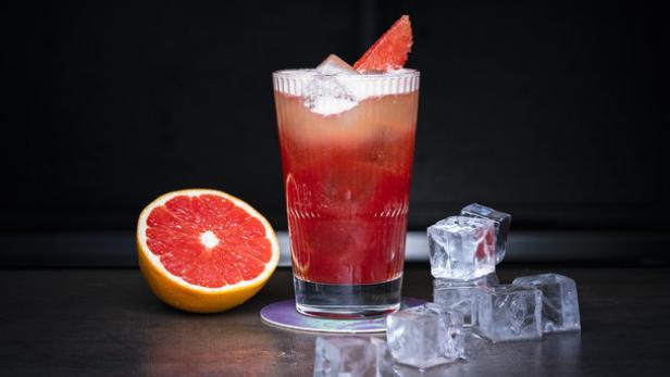 Ein Glas mit einem roten Cocktail, Eiswürfeln und einer halben Grapefruit auf einer Bar.