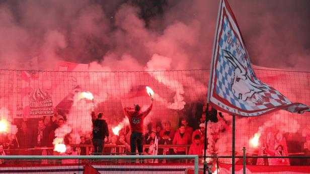 Fußballfans zünden rote Bengalos hinter einem Zaun mit einem Vereinsbanner.