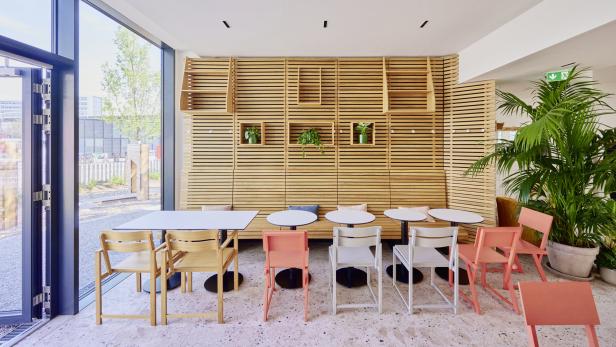 Ein heller Raum mit Tischen, Stühlen und einer hölzernen Wand mit Pflanzen.