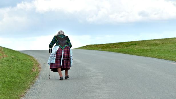 Eine ältere Frau mit Stock und traditioneller Kleidung geht allein eine Landstraße entlang.