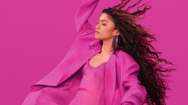Zendaya trägt einen pinkfarbenen Hosenanzug und eine passende Handtasche vor einem pinkfarbenen Hintergrund.