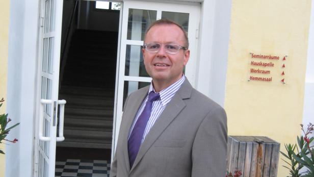 Ein Mann mit Brille und Anzug steht vor einem Gebäude mit der Aufschrift „Seminarräume“.