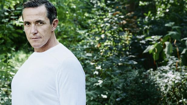 Ein Mann in einem weißen Hemd steht vor einem grünen Hintergrund.