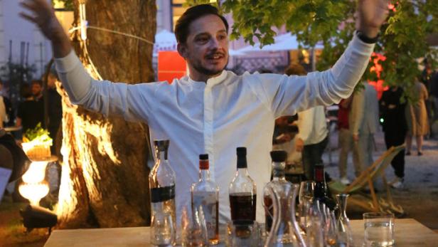 Ein Mann steht mit erhobenen Armen hinter einem Tisch mit Flaschen und Gläsern unter einem Baum mit Lichtern.