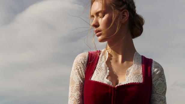 Eine Frau mit geschlossenen Augen trägt ein Dirndl vor einem bewölkten Himmel.
