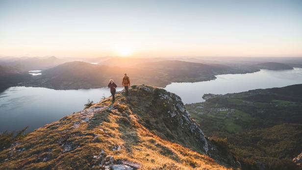 Zwei Wanderer auf einem Bergrücken mit Blick auf einen See bei Sonnenaufgang.