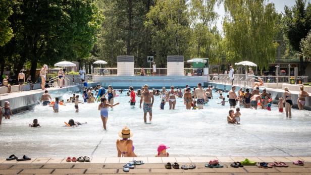 Viele Menschen, darunter Kinder und Erwachsene, genießen einen sonnigen Tag in einem Freibad mit flachem Wasser und umgebenden Bäumen.