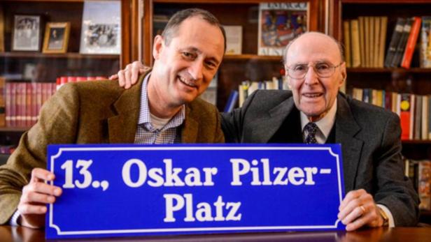 Zwei Männer halten ein blaues Schild mit der Aufschrift „13., Oskar Pilzer-Platz“.