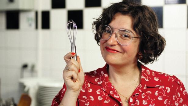 Eine Frau mit Brille hält einen Schneebesen und einen Topf in einer Küche.