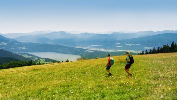 Zwei Wandererinnen auf einer blühenden Wiese mit Blick auf einen See und Berge.