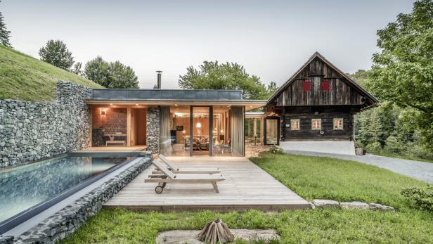 Ein modernes Haus mit Pool und ein traditionelles Holzhaus stehen nebeneinander.