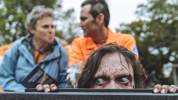 Ein blutverschmierter Zombie schaut über den Rand eines Behälters, während zwei Personen im Hintergrund stehen.