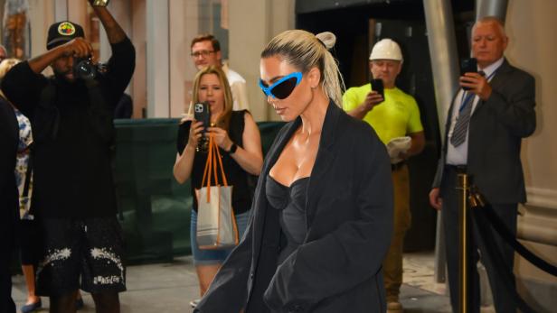 Kim Kardashian mit blauen futuristischen Sonnenbrille und schwarzem Blazer, umgeben von Fotografen.