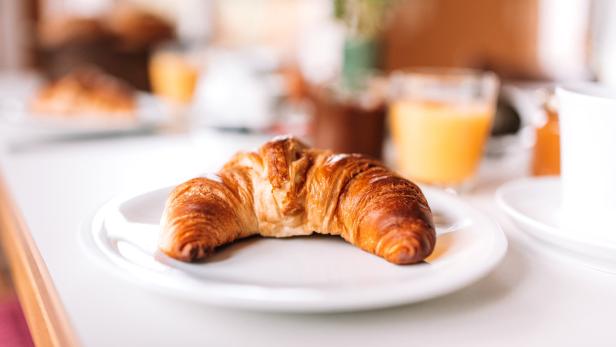 Ein Croissant liegt auf einem Teller, zusammen mit Orangensaft und Marmelade zum Frühstück.