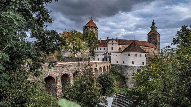 Burganlage mit Türmen, Brücke und Mauern über dichtem Grün unter düsterem Himmel.