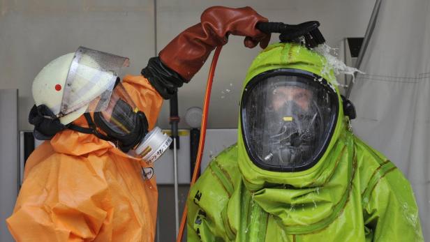 Eine Person in einem orangefarbenen Schutzanzug duscht eine andere Person in einem grünen Schutzanzug ab.