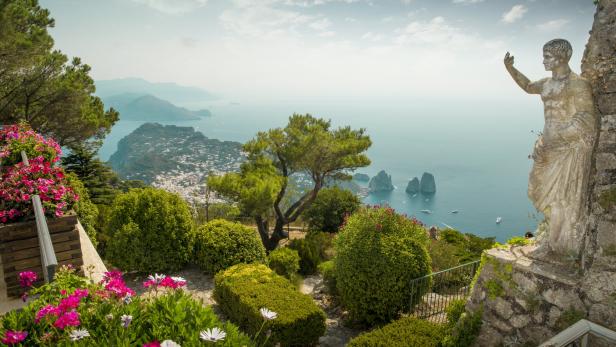 Blick von einem Garten auf Capri mit der Küstenlinie und einer Statue im Vordergrund.