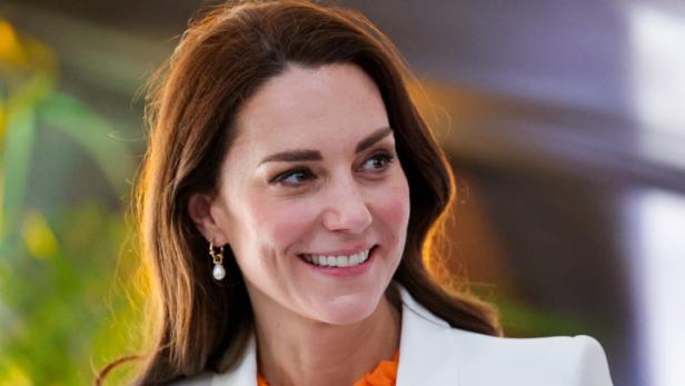 Kate Middleton lächelt in einem weißen Blazer und einer orangefarbenen Bluse.