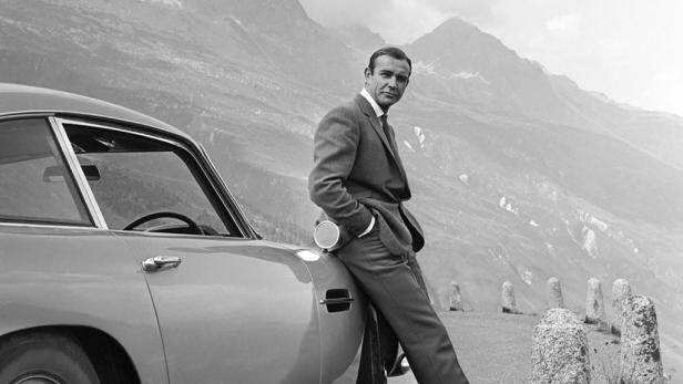 Sean Connery lehnt an einem Aston Martin vor einer Bergkulisse.