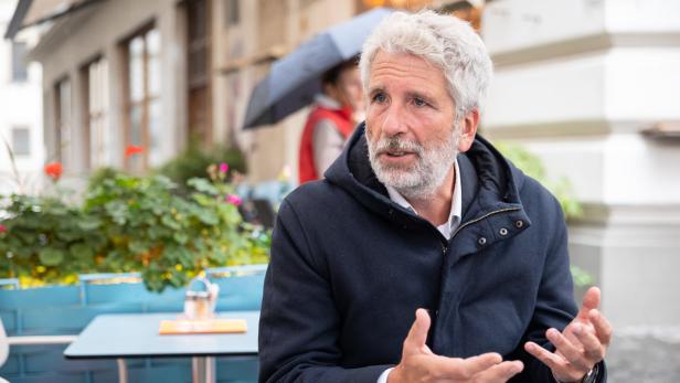 Ein Mann mit grauem Haar gestikuliert an einem Tisch im Freien.